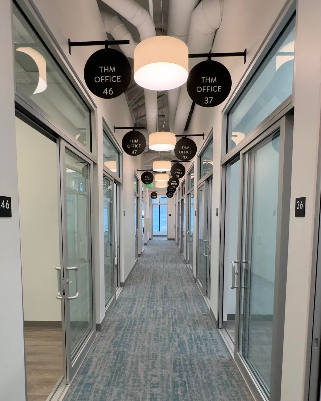 A long, modern hallway lined with individual **THM Office** suites, featuring glass doors and circular signs, showcasing the professional and contemporary workspace environment.