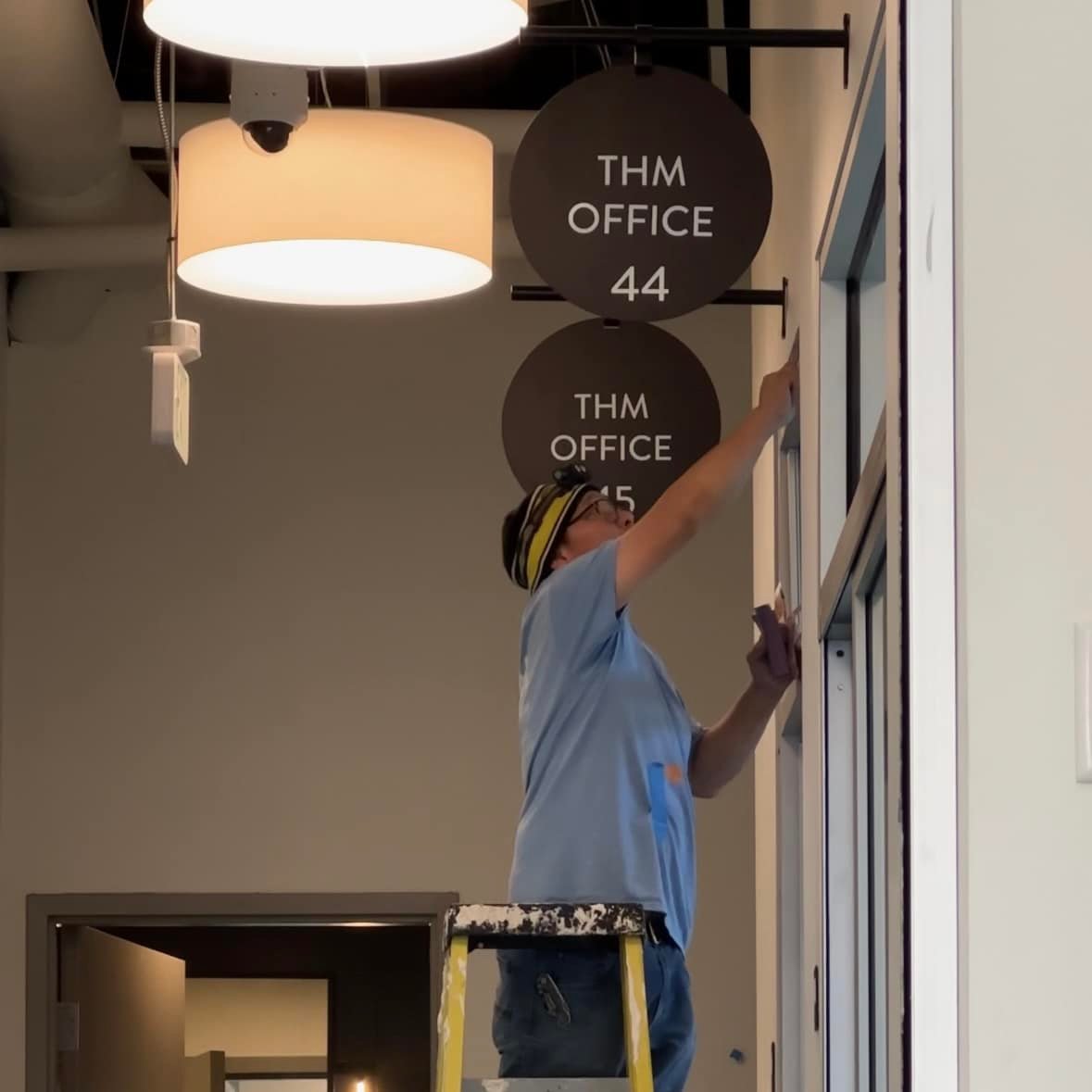 A worker installs directional signage for **THM Office** Suites, highlighting the final preparation of the office units for tenants.